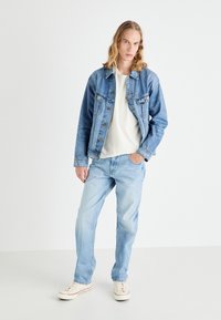 Jachetă din denim de culoare bleu deschis, cu închidere cu nasturi, buzunare pe piept și o croială lejeră, purtată cu blugi bleu deschis și adidași crem.