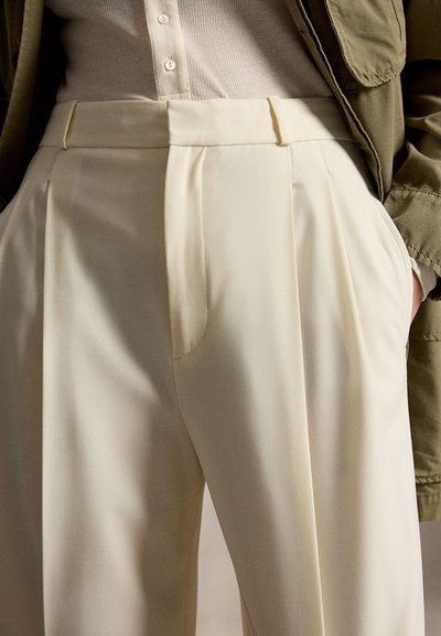 Προσαρμοσμένο παντελόνι στο χρώμα της κρέμας με πιέτες, κατασκευασμένο από απαλό ύφασμα. Διαθέτει επίπεδη μέση και πλαϊνές τσέπες.