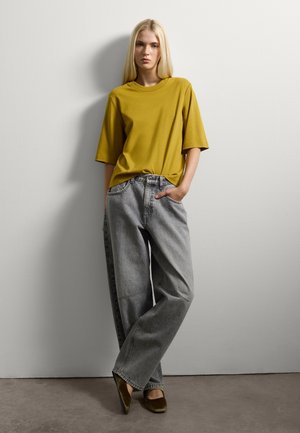 Donna con lunghi capelli biondi in piedi contro un muro bianco, indossa una maglietta larga color senape, jeans grigi larghi e ballerine marroni.
