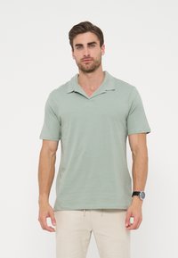 Άνδρας που φοράει ανοιχτό πράσινο πόλο με κοντά μανίκια και μπεζ παντελόνι, στέκεται μπροστά σε λευκό φόντο, με σκούρο ρολόι στον αριστερό καρπό.