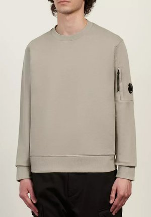 Uomo che indossa una felpa beige a maniche lunghe con una tasca con zip e un dettaglio circolare nero sul braccio sinistro, abbinata a pantaloni neri.
