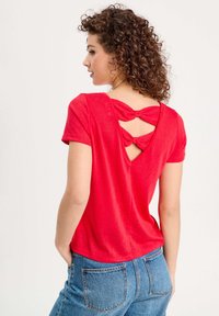 Rotes T-Shirt mit kurzen Ärmeln und V-Rücken-Design, verziert mit zwei Schleifen und einem strukturierten Stoff. Kombiniert mit hochtaillierten blauen Jeans.