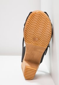 Semelle d'une chaussure à talon noire avec une semelle extérieure en caoutchouc texturé marron et un talon en bois, taille 37, appuyée contre un mur blanc.