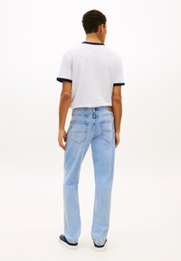 Mann som står med ryggen til, iført en hvit t-skjorte med mørk kant, lyseblå jeans og svarte joggesko med hvite såler.