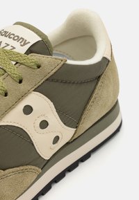 Zöld velúr és textil sportcipő krém színű részletekkel, kerek logóval, texturált felszínnel és fekete gumi talppal.