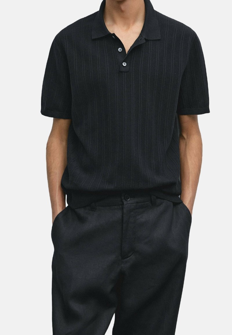 Homme portant un polo noir à manches courtes avec de fines rayures verticales et un pantalon noir, les mains dans les poches.