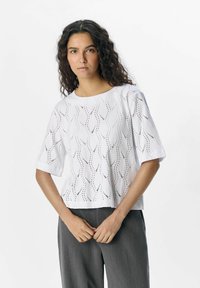 Haut blanc à manches courtes avec des détails en œillets à motifs ondulés. Texture lisse, encolure ronde et coupe décontractée. Assorti à un pantalon gris.