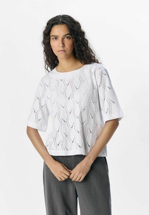 Haut blanc à manches courtes avec des détails en œillets à motifs ondulés. Texture lisse, encolure ronde et coupe décontractée. Assorti à un pantalon gris.