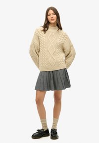 Beige strikket sweater med kabelmønster, høj hals; sort-hvid ternet plisseret nederdel; parret med sorte loafers og knæhøje strømper.