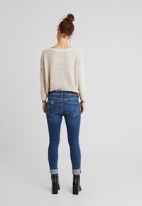 Neulepaita beigen värisenä, rento malli; siniset tiukat farkut, joissa on hieman kulutusta; mustat nilkkureissa, joissa on korot; näkyy takaa.