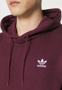 Mörkröd huvtröja tillverkad av mjukt tyg, med en vit broderad Adidas-logga på bröstet, försett med justerbara snören.