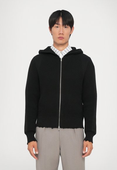 Sudadera con cremallera negra hecha de un tejido de punto texturizado. Presenta una capucha, puños de canalé y un dobladillo deshilachado, llevada sobre una camiseta a rayas.