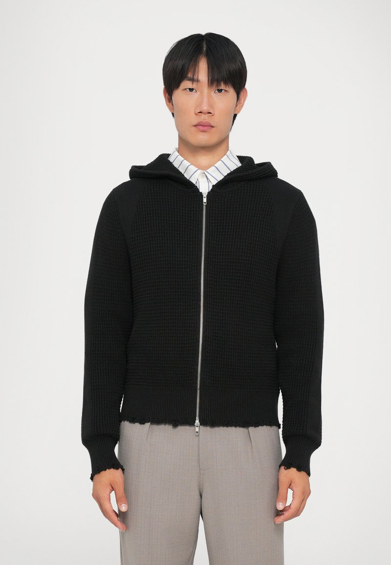 Svart zip-up hoodie gjord av texturerat stickat tyg. Har en huva, ribbade ärmslut och en fransad nederkant, bärs över en randig skjorta.