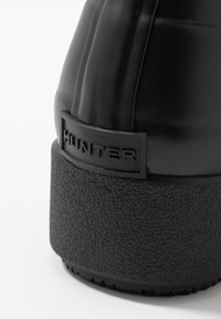 Botte en caoutchouc noir avec semelle noire texturée, arborant un logo « HUNTER » en relief sur un accent rectangulaire. Tige lisse avec des rainures verticales.