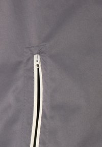 Gray fabric close-up featuring a zippered pocket with white trim. The texture appears smooth with stitched detailing around the zipper.