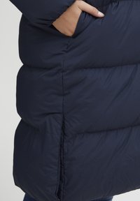 Mørkeblå pufferjakke med quiltet design, glatt tekstur og sidelommer, med en lett innsvinget midje for form.