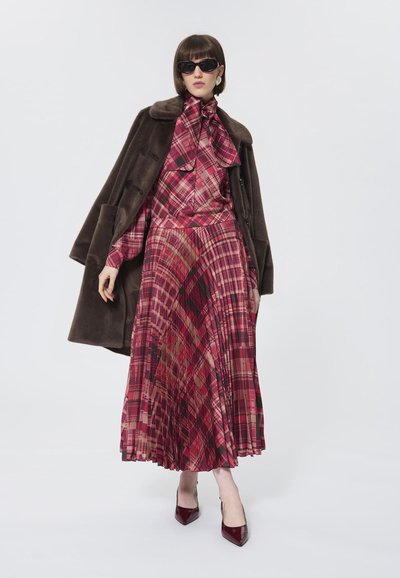 Abito lungo a pieghe in tartan rosso con blusa abbinata e colletto a fiocco, sovrapposto a un cappotto di pelliccia marrone scuro. Tacchi a spillo rossi.