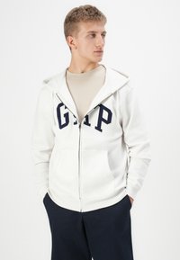 Hvit zip-up hettejakke laget av bomull, med et marineblått "GAP"-logo, frontlommer, justerbar hette og ribbede mansjetter. Båret over en beige skjorte.