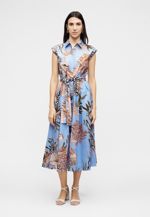 Femme portant une robe midi bleu sans manches avec des motifs floraux et coquillages orange et marron, nouée à la taille avec une ceinture en tissu assorti.