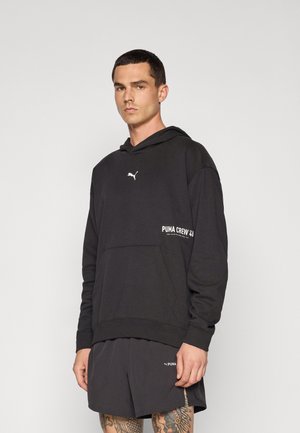Sort hættetrøje med hvidt Puma-logo og teksten "PUMA CREW 23," med en frontlomme og lange ærmer. Bæres med matchende shorts.
