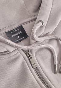 Svetlo šedá mikina na zips so soft textúrou, dekoračnými šnúrkami a strieborným zipsom. Na nášivke je uvedené "JOHNNY URBAN" a veľkosť "M."