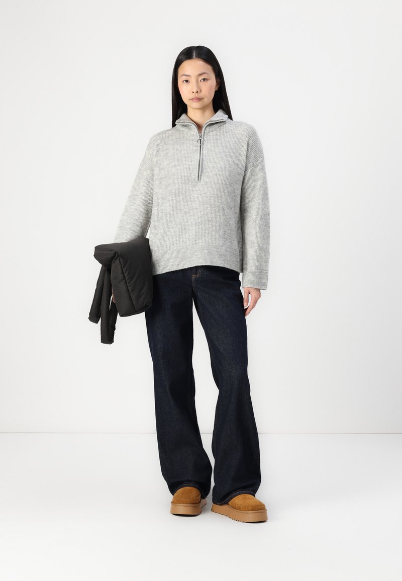 Szary sweter z dzianiny z kołnierzem na pół zamek, w połączeniu z ciemnymi dżinsami z szerokimi nogawkami i beżowymi butami wsuwanymi, trzymając czarną kurtkę puchową.