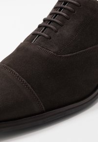 Chaussure en suède brun avec une finition lisse, bout rond et cinq trous de laçage ; présente des coutures subtiles et une semelle plate en caoutchouc.