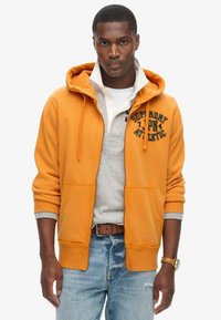 Felpa arancione con zip e dettagli grigi, dotata di tasca frontale e ricamo "Superdry Athletic". Abbinata a un maglione grigio chiaro e jeans.