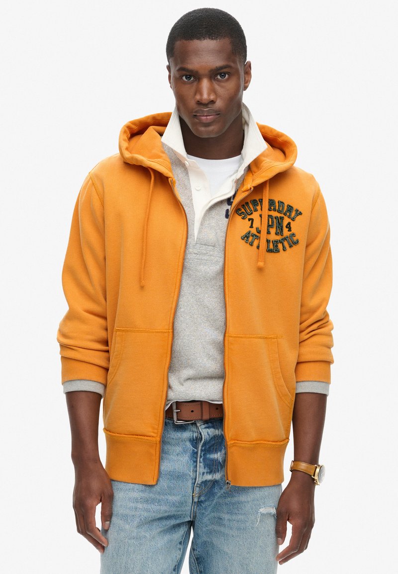 Felpa arancione con zip e dettagli grigi, dotata di tasca frontale e ricamo "Superdry Athletic". Abbinata a un maglione grigio chiaro e jeans.