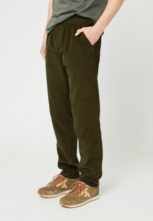 Persoon in olijfgroene corduroy broek met elastische tailleband en tan sneakers met oranje veters, staand tegen een effen achtergrond.