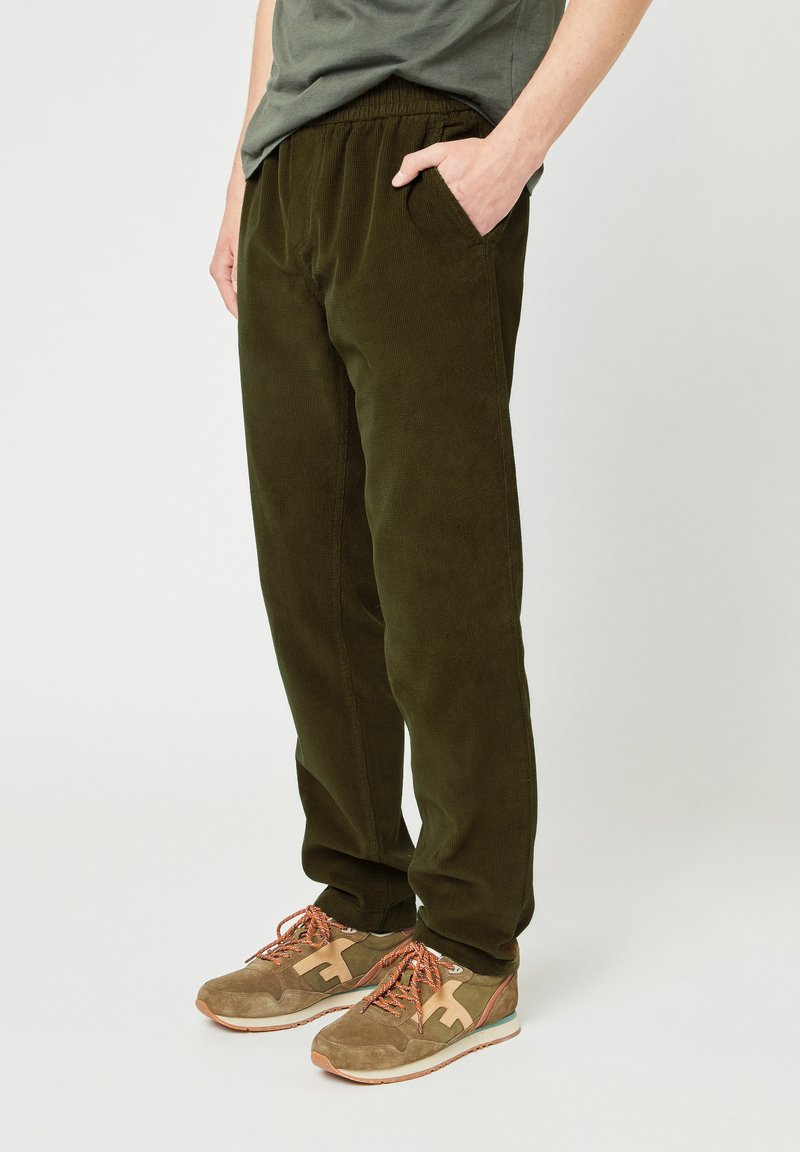 Persoon in olijfgroene corduroy broek met elastische tailleband en tan sneakers met oranje veters, staand tegen een effen achtergrond.