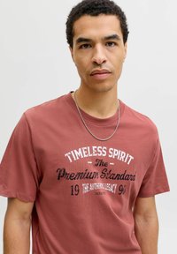 Joven con una camiseta de color óxido que lleva el texto "Timeless Spirit Premium Standard 1990" y una cadena de plata, sobre un fondo liso.