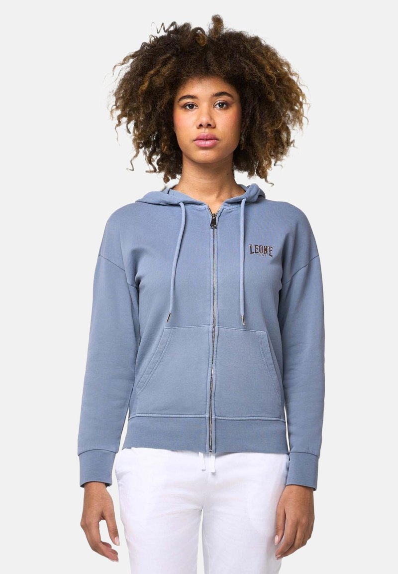 Femme aux cheveux bouclés portant un sweat à capuche bleu clair zippé avec des poches devant et un pantalon blanc, debout devant un fond clair uni.