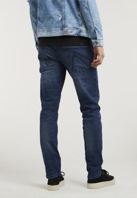 Jeans de color azul oscuro con un corte slim, que cuentan con bolsillos traseros y un ligero desgaste. Combinados con una camisa negra y zapatos de ante negros.