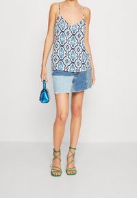 Žena v bezramínkovém vzorovaném topu, patchworkovém denimovém mini sukni, zelených sandálech s tkaničkami, držící malou kovovou modrou kabelku.