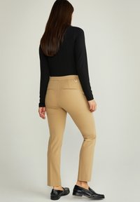 Beżowe spodnie o dopasowanym kroju z gładką fakturą, posiadające dwie tylne kieszenie oraz okrąły akcent. Noszone z dopasowaną, czarną bluzką z długim rękawem i czarnymi mokasynami.
