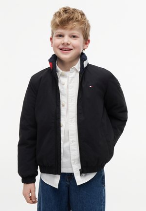 Lächelnder Junge mit kurzen blonden Haaren, der eine schwarze Jacke mit Reißverschluss über einem weißen Hemd mit Knöpfen und blauen Jeans trägt und vor einem schlichten weißen Hintergrund steht.
