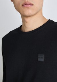 Black long-sleeve shirt featuring a round neckline and a small black leather logo patch on the left chest area. Smooth, soft texture.
