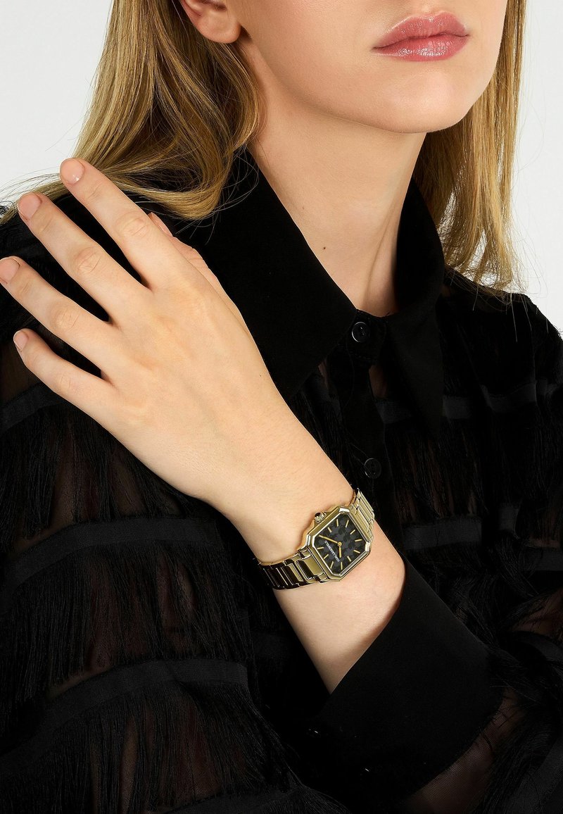 Goudkleurig octagonaal horloge met een zwart wijzerplaat, slanke armband en contrasterend zwart overhemd met franjes.