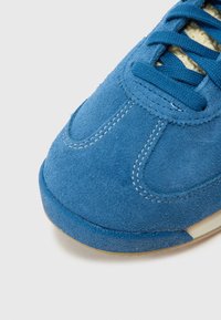 Mėlynas zomšiniai sportbačiai su balta siūle, apvalia nosimi ir tekstūruota paviršiaus. Turi derančias raištelius ir kontrastingą smėlio spalvos pado detalę.