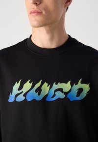 Male torso wearing a black crew neck sweatshirt with "HUGO" in green-to-blue flame-style lettering across the chest.