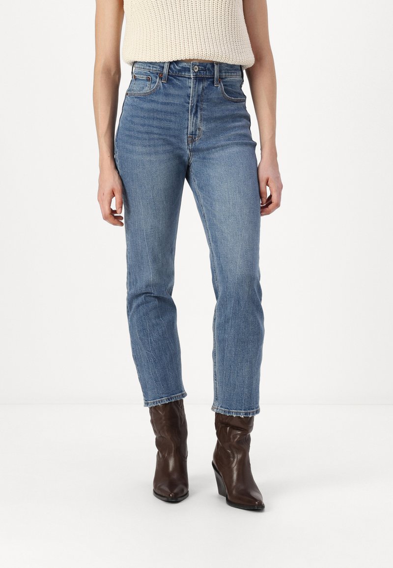 Jeans en denim bleu taille haute avec une coupe droite, présentant un délavage et un design classique à cinq poches. Associés à des bottes à talons marron.