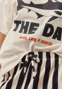 Witte t-shirt met grote zwarte tekst en haai afbeelding; gecombineerd met marineblauwe en witte gestreepte shorts met een trekkoord taille.