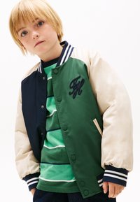 Blondes Kind mit Sommersprossen, das eine grün-navy-hellbeige College-Jacke über einem grün gestreiften Hemd trägt und vor einem weißen Hintergrund steht.