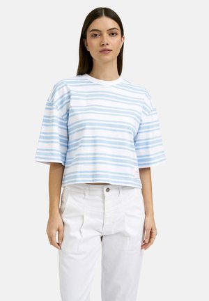 Woman wearing a loose, short-sleeve white and light blue striped shirt with white high-waisted pants, standing against a plain background.