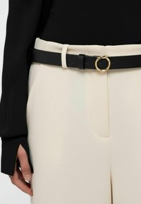 Černý kožený pásek se zlatou kulatou sponou, nošený přes krémové kalhoty. Hladká textura, minimalistický design, viditelné poutka na pásek.