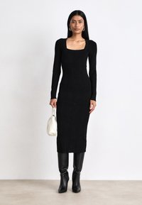 Black knitted long-sleeve dress with a square neckline, form-fitting shape, vertical ribbed texture, paired with knee-high black boots and a white handbag.