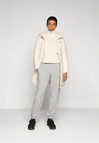 Biela vatovaná bunda s vysokým golierom cez svetlý sveter, v kombinácii so sivými teplákmi a čiernymi športovými topánkami. Minimalistický dizajn.