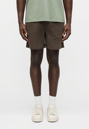 Homme portant un short marron, une chemise vert clair, des chaussettes blanches et des baskets blanc cassé, debout devant un fond blanc uni.