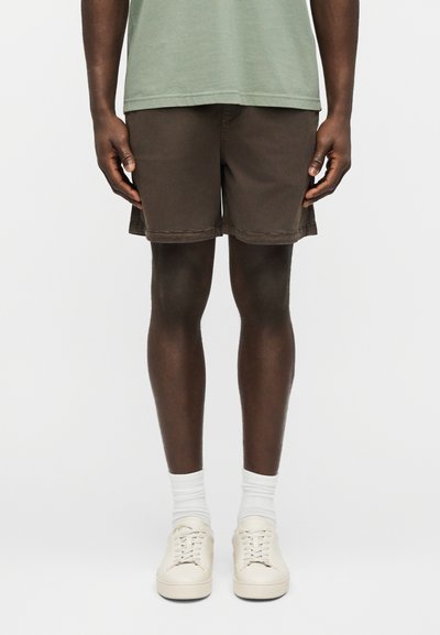Homme portant un short marron, une chemise vert clair, des chaussettes blanches et des baskets blanc cassé, debout devant un fond blanc uni.